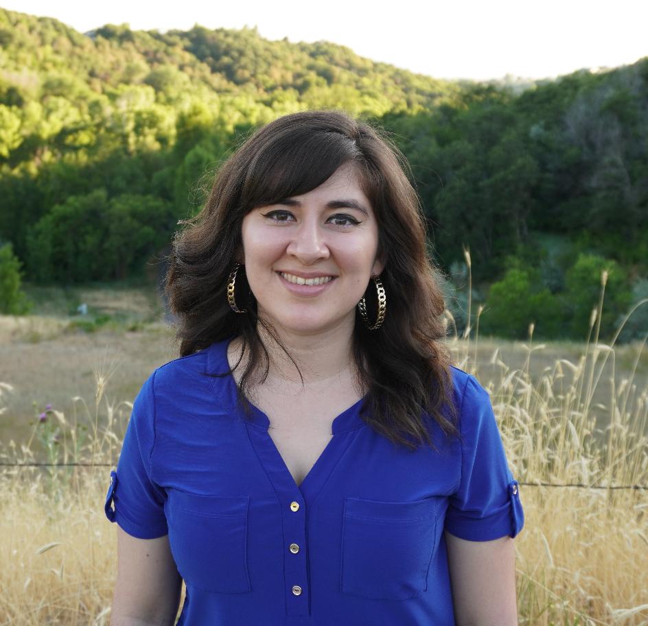 Kaela Rivera stands in front of a barbed wire fence with desert grasses behind her and rolling green mountains. She wears a blue shirt and smiles as her gold hoops earrings catch the evening light. 